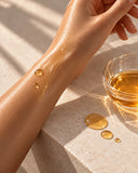 Kanya body oil glistening on the skin of a forearm, with a glass dish of oil on travertine